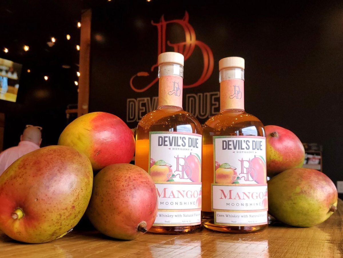 Apple, Fruit, Baby, Person. Text: DUE DEV DEVIL&#039;S DUE DEVIL&#039;S DUE MANGO MANGO MOONSHINE MOONSHINE Whiskey with Natural Flavors