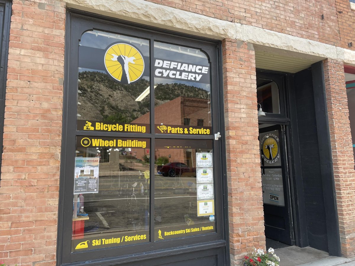 Brick, Postal Office, Shop, Person. Text: DEFIANCE CYCLERY Bicycle Fitting Parts &amp; Service Wheel Building RIDES&amp; REGGAE Hours defiancecyclery.com 970-984-7022 OPEN ring Backcountry Ski Sales/Rentals Ski Tuning/Services